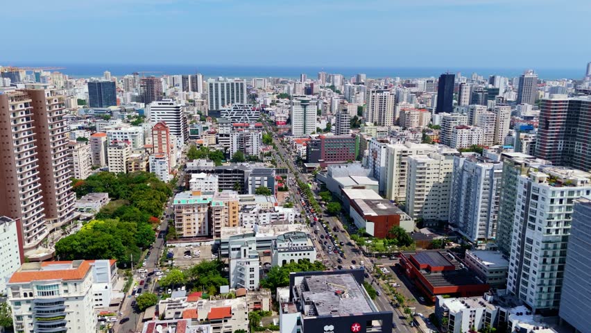 Dense high-rise towers line straight boulevard busy with traffic, Caribbean Sea visible past coastal condos beneath clear sunny sky in Santo Domingo, Dominican Republic. Aerial drone wide shot