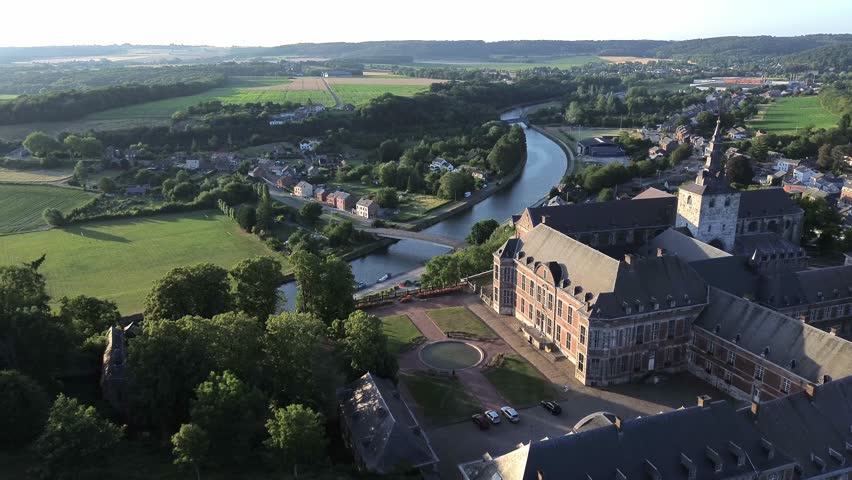 Aerial view captures historic Floreffe Abbey in Namur, Belgium. River Sambre winds gracefully through green fields and village homes. Stone buildings with dark roofs sit near tree-lined paths