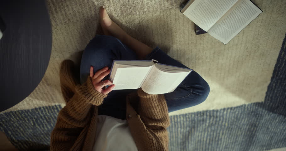 Calm Home Study Moment as a Young Woman Reads a Book on a Living Room Rug. Creative Overhead-to-wide Camera Movement Capturing Focused Leisure and Mindful Learning at Home.