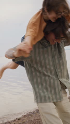 Vertical video of Caucasian father carrying young son on shoulders, spinning and laughing on beach. Footage is ideal for family travel campaigns and parenting lifestyle advertisements