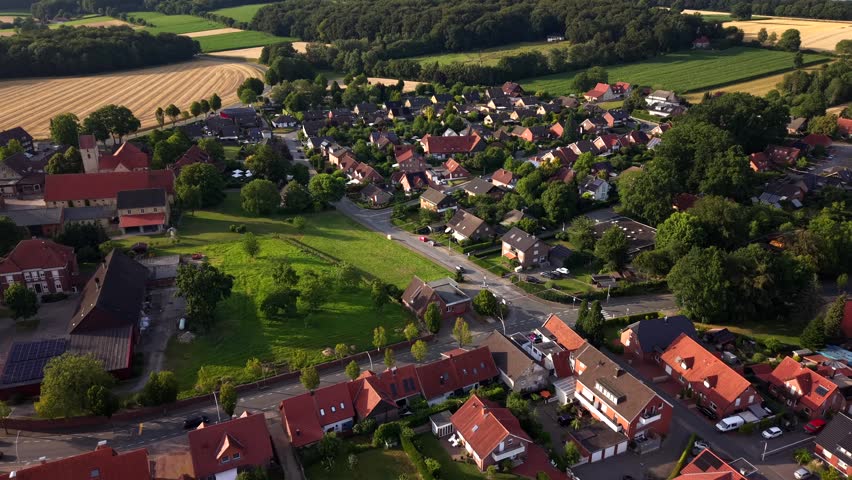 Small village surrounds historic church steeple near Dülmen North Rhine-Westphalia Germany, golden wheat fields and farmland stretch beyond red-tile rooftops at sunset. Aerial drone flyover shot