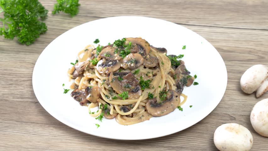 Close-up of a hand garnishing creamy mushroom spaghetti with fresh parsley on a white plate. Homemade Italian pasta with sautéed mushrooms in a rich cream sauce, captured in a bright kitchen setting. Perfect for food, cooking, restaurant, and recipe concepts.
