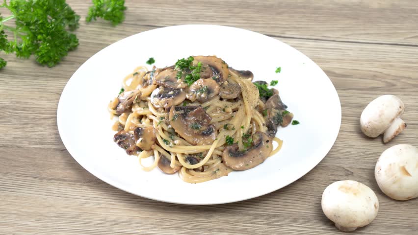 Close-up of a hand garnishing creamy mushroom spaghetti with fresh parsley on a white plate. Homemade Italian pasta with sautéed mushrooms in a rich cream sauce, captured in a bright kitchen setting. Perfect for food, cooking, restaurant, and recipe concepts.