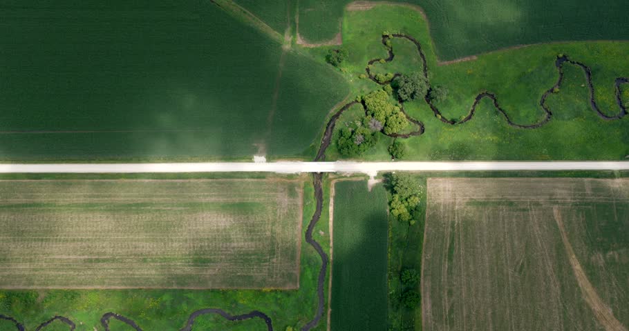 Meandering stream winds through cultivated farmland beneath a dirt farm road, revealing riparian vegetation, geometric field patterns and drifting cloud shadows, top down drone ascent.