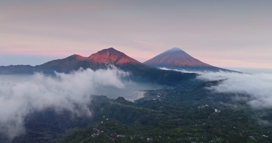 Bali volcanic landscape featuring Mount Batur and Mount Agung volcanoes towering above Lake Batur, surrounded by low morning clouds and lush green forests at a soft sunrise light. Aerial drone flight