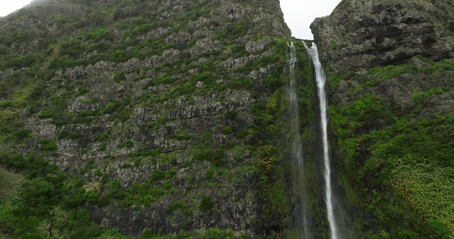 A drone view of Poco do Bacalhau Waterfall plunges down a narrow gap between two foggy steep, green-covered mountainsides in Flores island, Azores