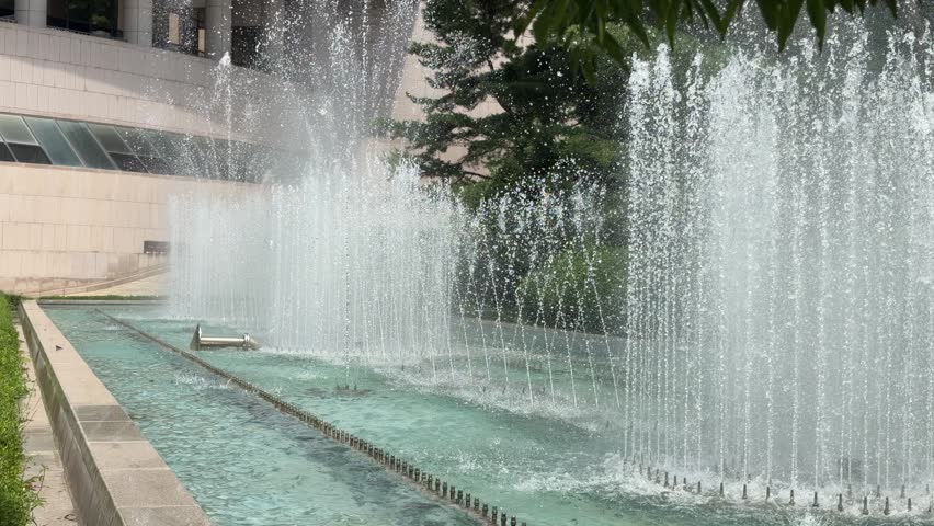 Diagonal view along a long musical fountain pool with dense rows of water jets spraying beside a stone terraced wall on a bright summer day in Seoul, South Korea