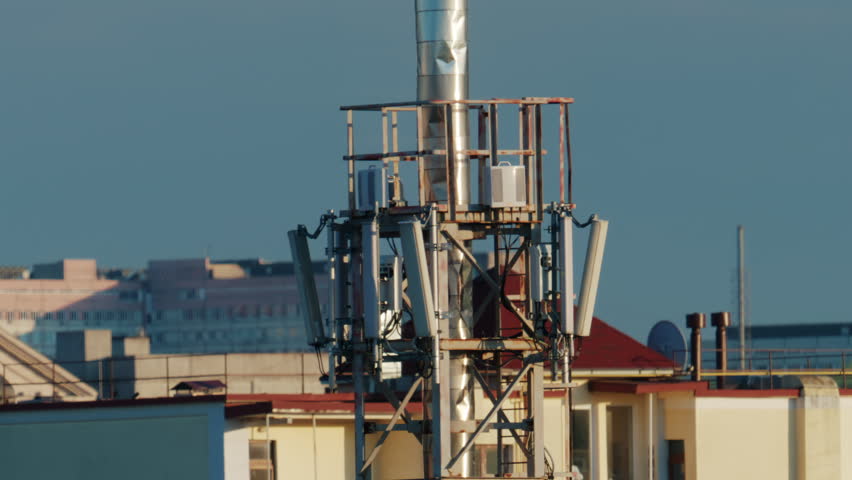 Urban rooftop scene featuring a cellular communication tower with multiple panel antennas, mounted on a metal framework above residential buildings