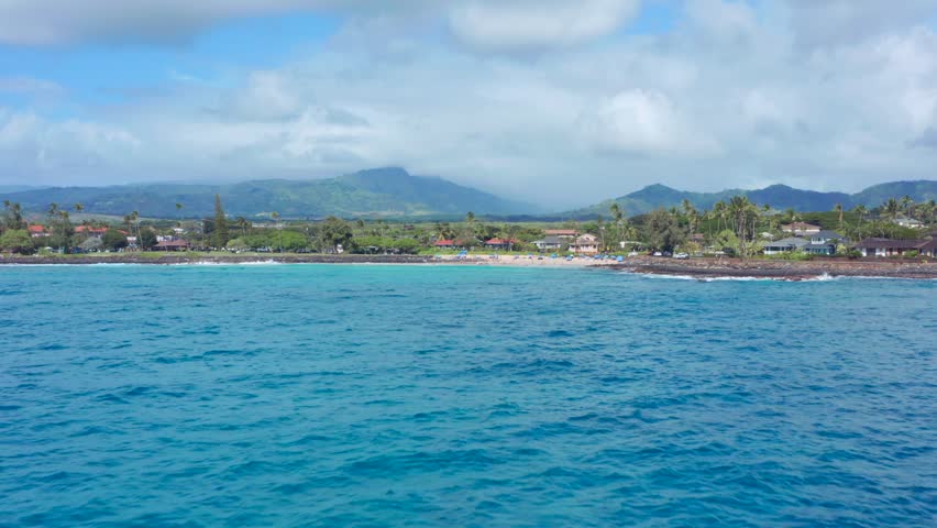 Picturesque coastline of Poipu, Kauai, where crystal-clear turquoise waters meet golden beaches, lush tropical landscapes, and swaying palm trees beneath dramatic island mountains. This peaceful ocean perspective captures the timeless beauty of Hawaii, making it perfect for travel campaigns, destination marketing, tourism, luxury resorts, nature films, and premium coastal stock footage. fast aerial drone flight over water surface