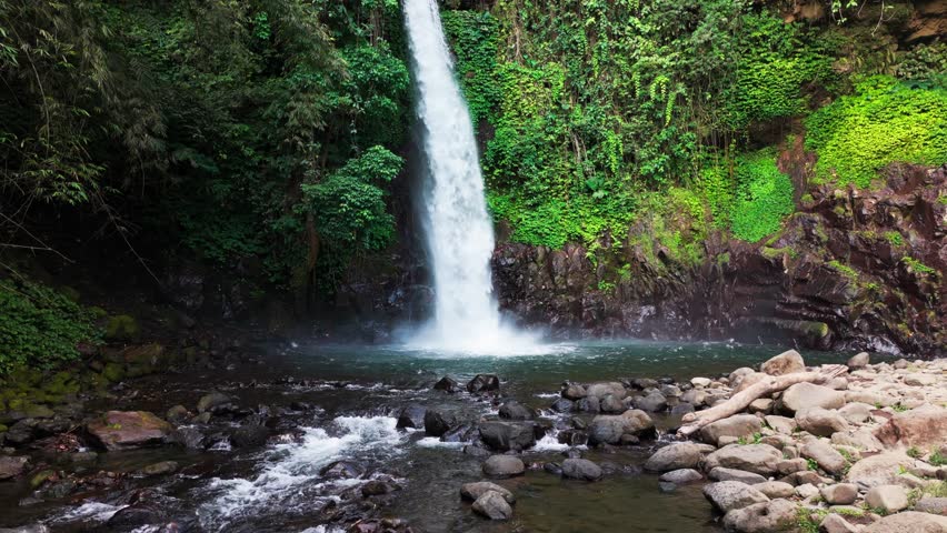 Aerial view of a waterfall cascading through basalt columns. Water plunges into a deep, narrow gorge. Rugged, jagged cliffs frame the falling stream. Exposed earth and stone surround the dramatic drop