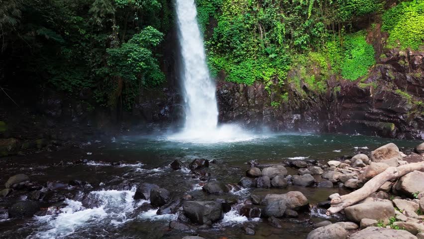 Aerial view of a waterfall cascading through basalt columns. Water plunges into a deep, narrow gorge. Rugged, jagged cliffs frame the falling stream. Exposed earth and stone surround the dramatic drop
