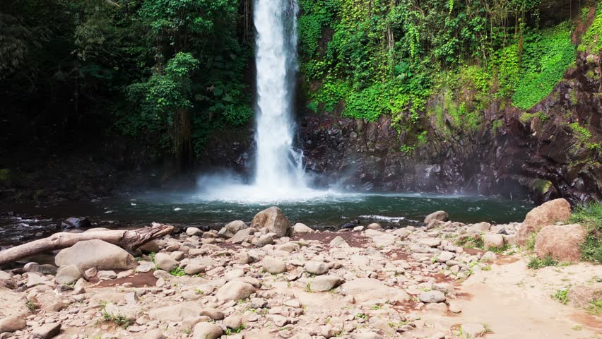 Aerial view of a waterfall cascading through basalt columns. Water plunges into a deep, narrow gorge. Rugged, jagged cliffs frame the falling stream. Exposed earth and stone surround the dramatic drop