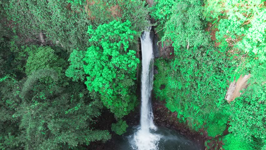 Aerial view of a waterfall cascading through basalt columns. Water plunges into a deep, narrow gorge. Rugged, jagged cliffs frame the falling stream. Exposed earth and stone surround the dramatic drop