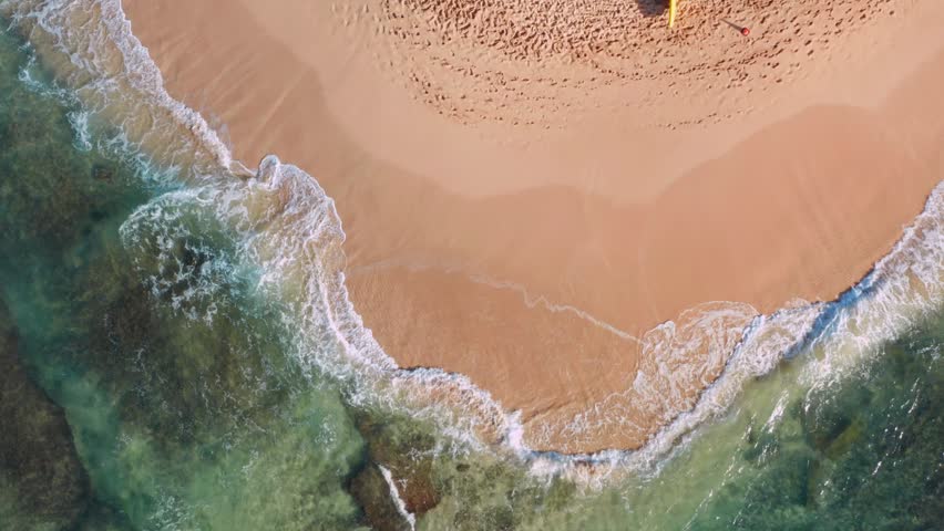 Aerial drone shot rising over Poipu Beach Park on Kauai, Hawaii, revealing waves rolling onto a pristine golden sandy beach from both the left and right sides. Crystal-clear turquoise water, a prominent lifeguard tower, and the picturesque tropical coastline create a breathtaking island scene, perfect for showcasing Hawaii travel, beach vacations, coastal landscapes, ocean beauty, surfing culture, and tropical paradise destinations.