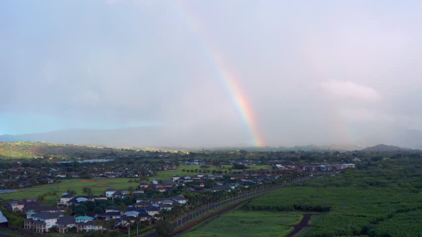 Aerial drone view of a Hawaii residential neighborhood beneath a vibrant rainbow after rainfall, showcasing lush green landscapes, tropical scenery, and peaceful island living.