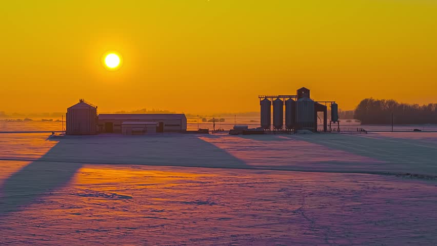 Aerial view of farm silos in a snow-covered field during a vibrant golden sunset in Latvia.