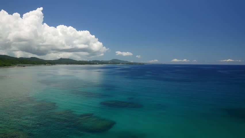 A drone view of calm deep-blue coastal waters with turquoise shallows, distant green hills, and bright skies in Okinawa, Japan