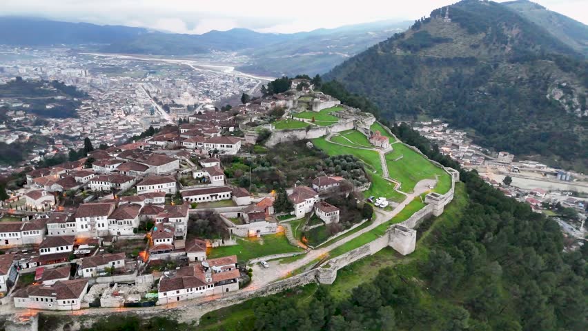 High-angle aerial shot circling the ancient stone fortification walls, traditional residential quarters, green lawns, and hilltop Holy Trinity Church inside Berat Castle, Albania