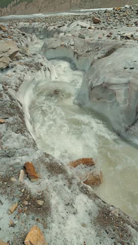 Rushing glacial river carves path through icy canyon surrounded by rocky terrain. Muddy water flows over ice stone under natural daylight. Composition highlights erosion raw mountain environment