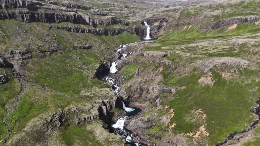 Aerial view of a cascading waterfall flowing through a rugged, green mountain valley in Iceland.