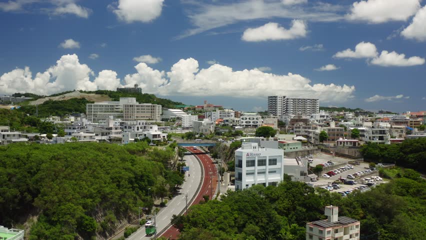 A drone view of a hillside cityscape with modern buildings, lush green trees, and winding red-toned road under bright skies in Okinawa, Naha, Japan