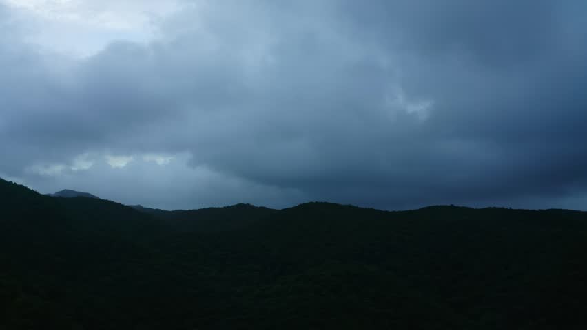 A wide-angle shot of heavy storm clouds hanging above forested hills, low light and dark tones conveying dramatic Okinawa weather