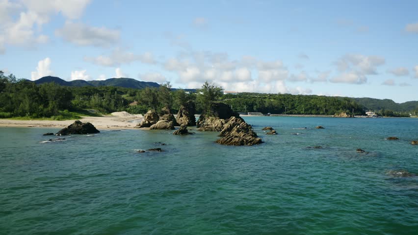 A frontal view of rocky formations emerging from shallow coastal water with green hills behind in Okinawa, Japan