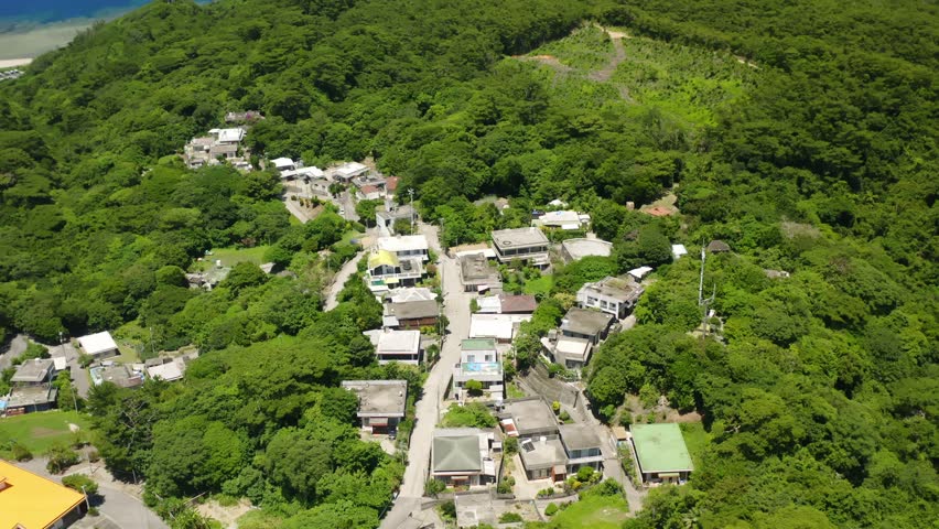 A drone view of a small residential area surrounded by dense forested hills, highlighting rural life in Okinawa, Japan