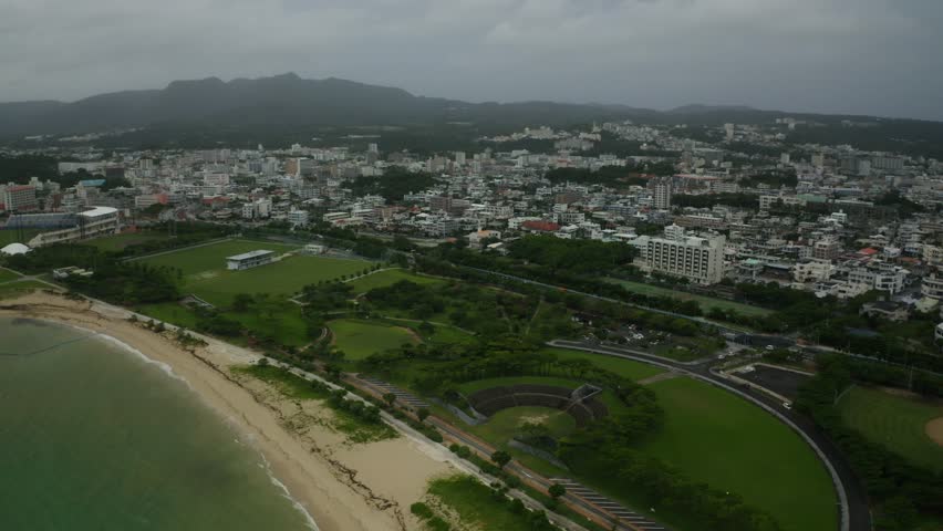 An Aerial view of the coastal Nago city with sandy beaches, green parks, and distant mountains beneath cloudy skies in Okinawa, Japan