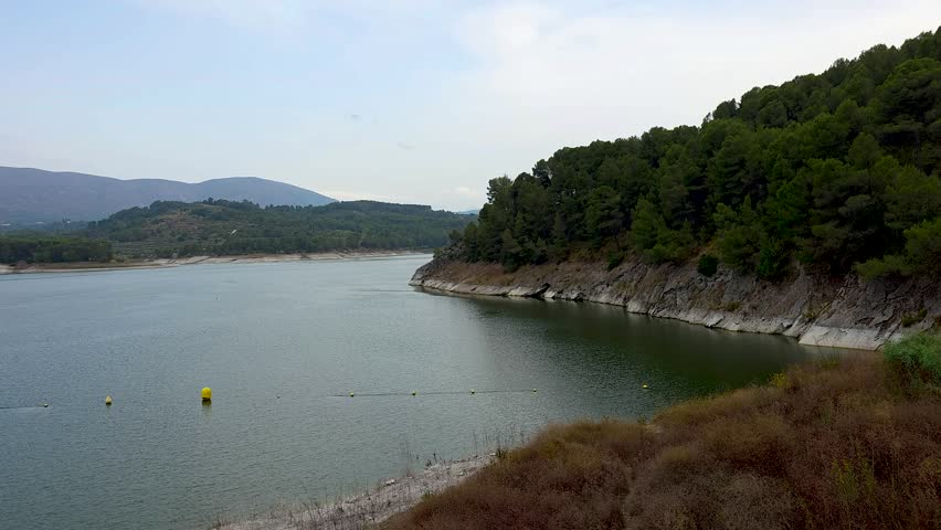 View of Beniarres reservoir lake from dam bridge with yellow safety buoys line, panoramic mountain range and serene water surface, Alicante, Spain.
