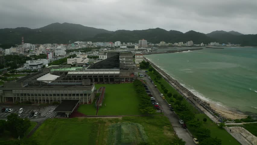 An Aerial view of a coastal cityscape with oceanside roads, modern buildings and misty mountains under cloudy sky in Nago, Okinawa, Japan