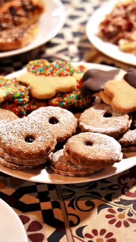 Sweet cookies pile high, dusted with sugar. Gingerbread shapes, colorful, chocolate-dipped, stacked neatly. Warm, inviting spread, set upon patterned tablecloth. Ideal for holiday, bakery