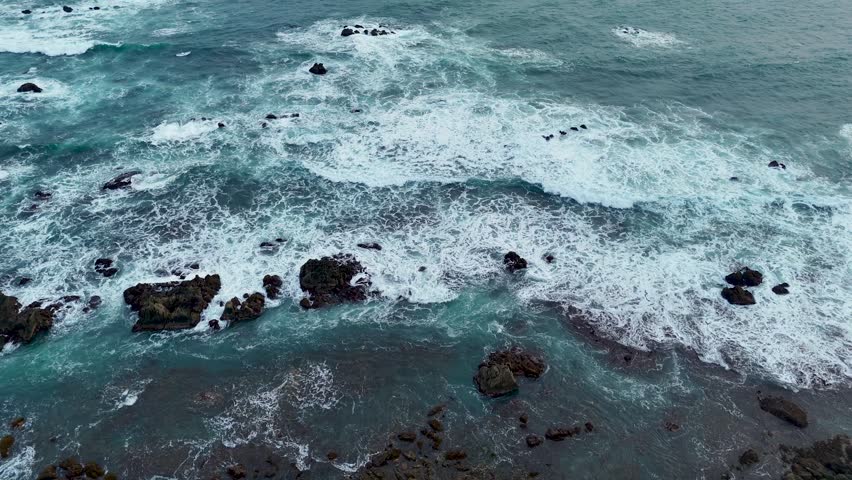 Aerial view of turquoise ocean waves crashing against dark volcanic rocks along the rugged coastline of Indonesia.