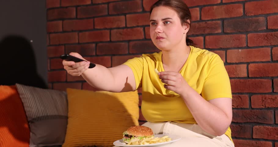 Overweight woman eating French fries and watching TV on sofa near red brick wall at home