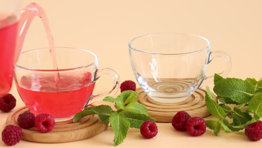Pouting tasty raspberry tea from teapot into glass cups, fresh berries and mint leaves on beige background, closeup