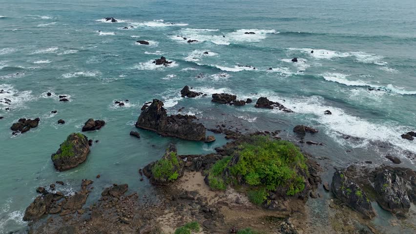 Aerial view of rocky coastline with crashing waves and lush green vegetation on volcanic rock formations along the coast of Indonesia.