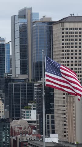 American flag ripples before downtown Seattle skyscrapers, glass and steel towers rising beneath overcast gray clouds, dense urban high-rise district framing city center. Aerial drone vertical orbit