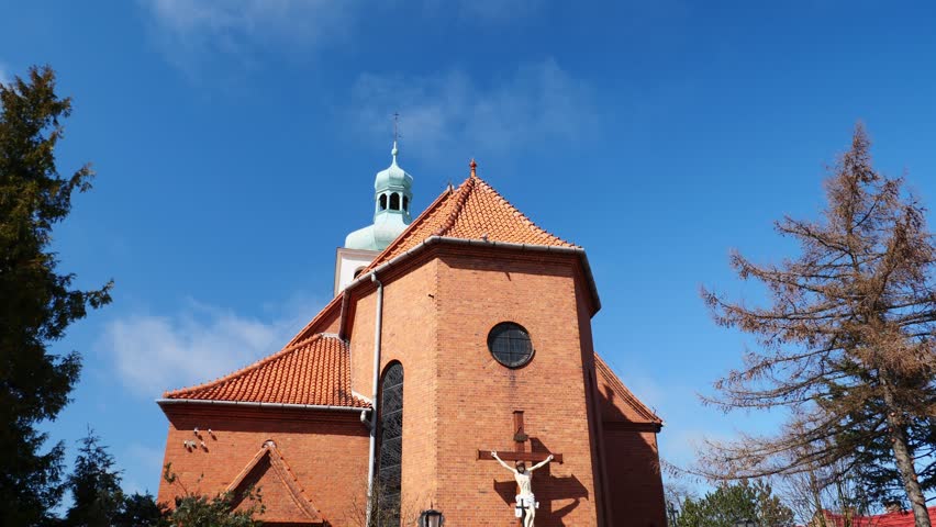 A graceful tilt-down shot reveals the red-roofed Church of the Visitation of the Blessed Virgin Mary in Jastarnia, Poland, highlighting its historic architecture against the coastal sky.