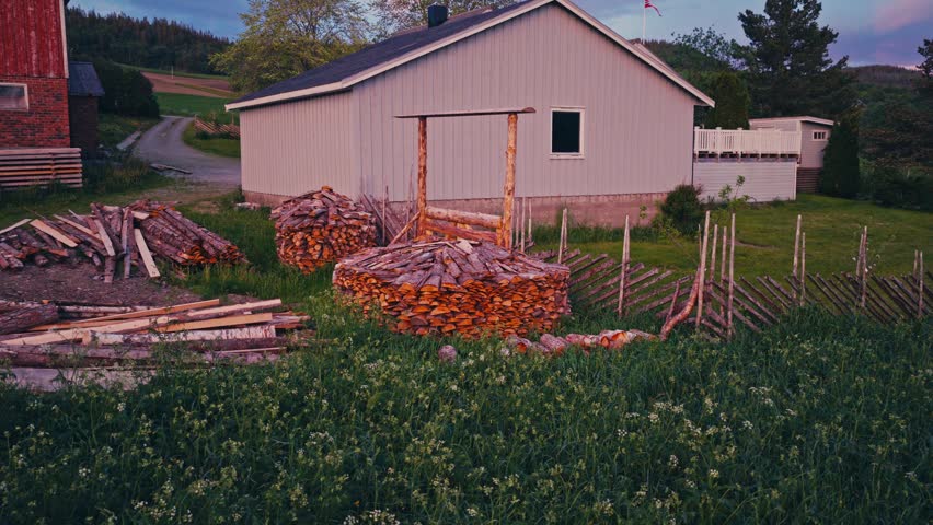 A Pile Of Logs Stands Beside A Wooden Barn And House In A Rural Countryside Landscape. Aerial Rotate Shot