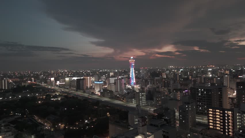 Aerial view of the illuminated Tsutenkaku Tower standing amidst the dense urban skyline of Osaka at dusk, with city lights glowing across the streets of Osaka, Osaka, Japan.