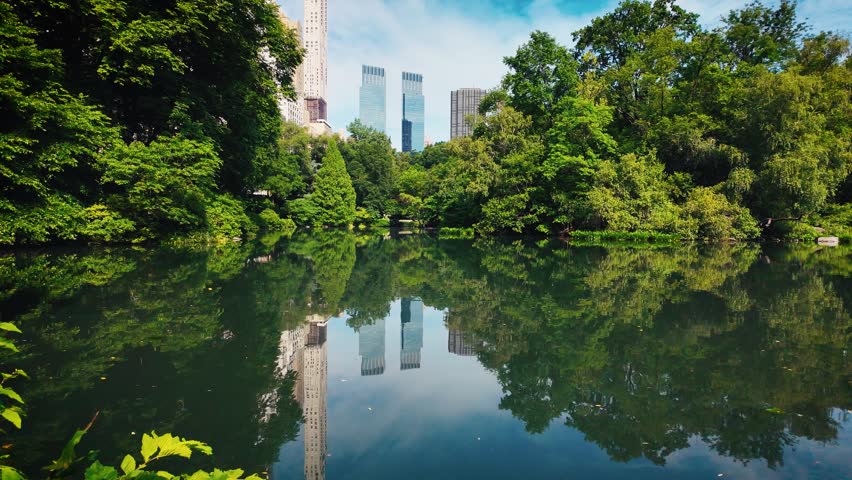 Beautiful view across Central Park Lake with calm reflections, lush green trees and modern Manhattan skyscrapers creating a peaceful urban nature scene in New York City.