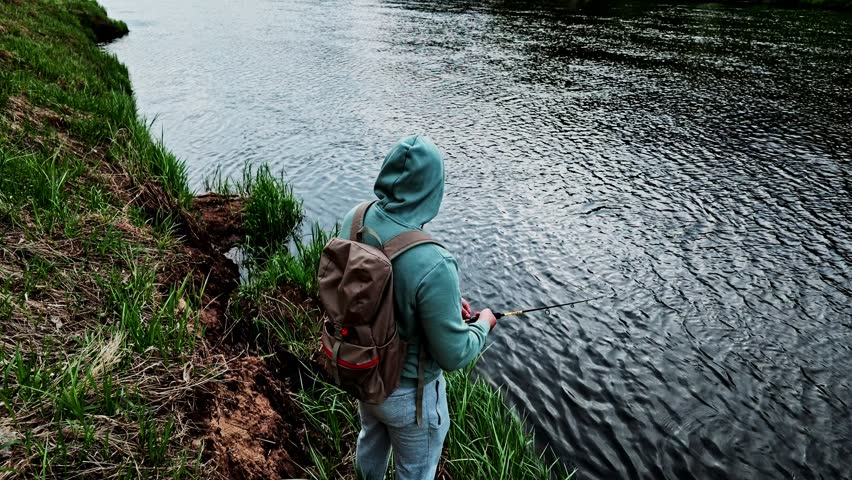 Fisherman in a hoodie holding a spinning rod and fishing from the grassy bank of a river