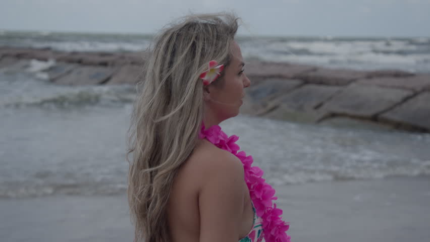 A smiling girl with a flower in her hair and pink lei walks along Galveston Beach at sunset, waves crashing on rocks in the soft evening light.