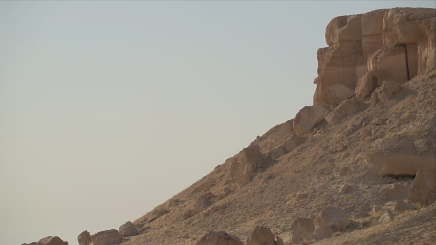 A barren desert rock hill rises against a pale sky in a vast, empty arid landscape.