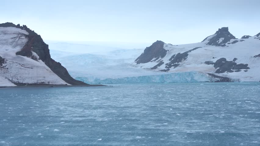 Cruising Antarctica Glacier Cloudy Day with Floating Ice Bergs Snow Covered Mountains. Rocky Beach. Blue Frozen Waters View From Cruise Ship.