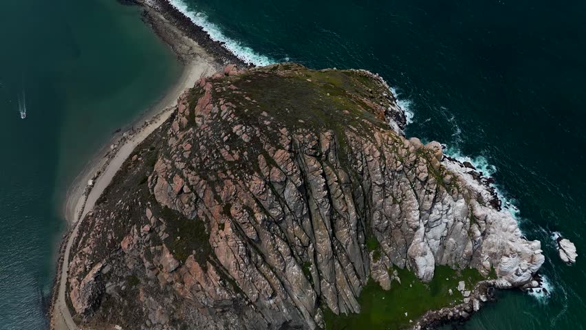Aerial view of the rugged volcanic Morro Rock formation surrounded by the Pacific Ocean and a sandy spit in Morro Bay, California, United States.