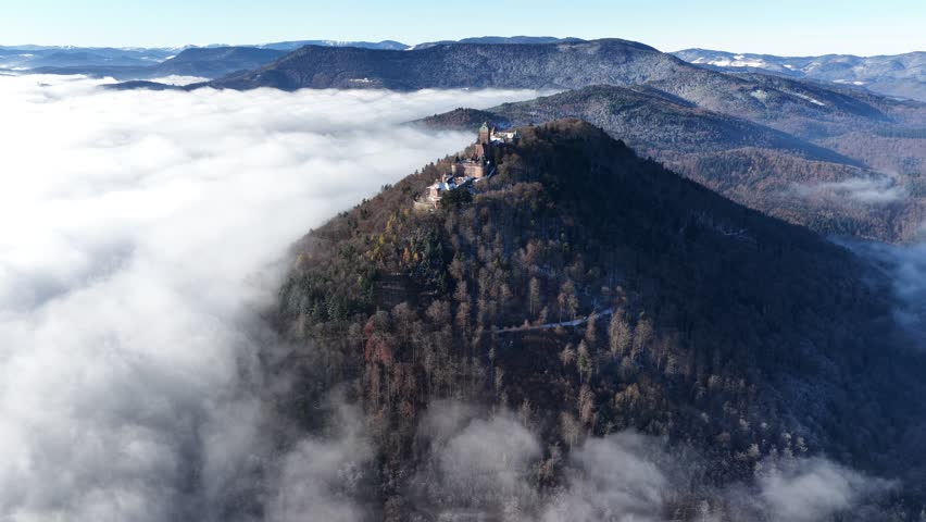 A drone view of Chateau du Haut-Koenigsbourg perched on its forested mountain, rising above swirling fog with distant ridges forming a layered horizon