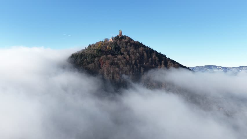 An aerial view of Chateau du Haut-Koenigsbourg rising through thick mountain fog, its wooded slopes glowing under the clear blue sky above
