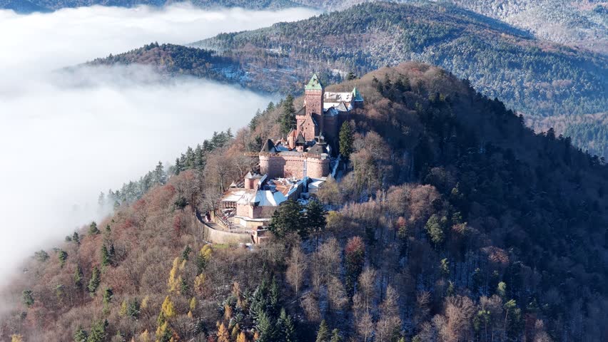 A drone view of Chateau du Haut-Koenigsbourg rising above a forested mountain ridge, soft clouds filling the Alsace valley below in morning light