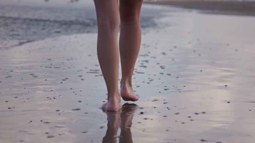 Close-up tracking shot of a joyful girl walking barefoot along Galveston Beach at sunset, gentle waves reflecting pastel skies on wet sand.
